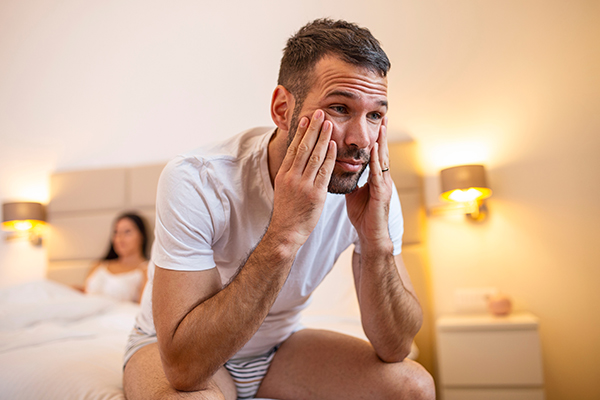 Discouraged man sitting on edge of bed with female partner in the background
