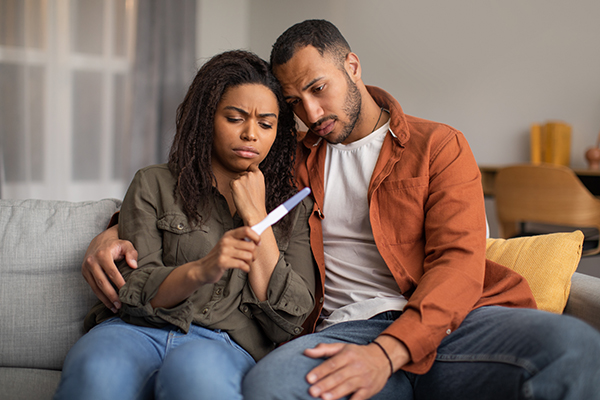 couple sitting on couch looking disappointed at pregnancy test held by woman
